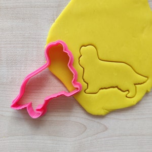 Może przedstawiać: Różowa plastikowa foremka do ciasteczek w kształcie psa rasy Cavalier King Charles Spaniel. Foremka leży na żółtej plastelinie, która została wycięta w tym samym kształcie.