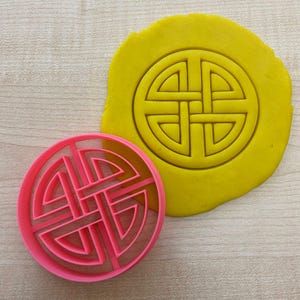 May include: A pink plastic cookie cutter in the shape of a Celtic knot design. The cutter is shown on top of a yellow piece of modeling clay with the same design imprinted in the clay.