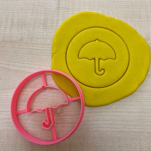 Pode incluir: Um cortador de biscoitos de plástico rosa em forma de guarda-chuva. O cortador está sobre uma superfície amarela com uma impressão do guarda-chuva.