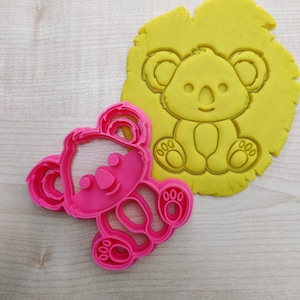 Op de afbeelding: Een roze plastic koala-koekjessnijder naast een gele koala-vorm in deeg gedrukt. De koekjessnijder heeft de vorm van een koala met een lachend gezicht, ronde oren en drietenige voeten.