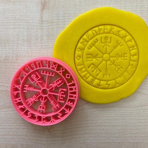 Peut inclure: Un emporte-pièce en plastique rose en forme de boussole circulaire avec des runes autour du bord. La boussole a quatre points cardinaux avec des symboles et un soleil au centre. L'emporte-pièce est sur une surface en bois à côté d'une empreinte jaune de l'emporte-pièce dans de la pâte.