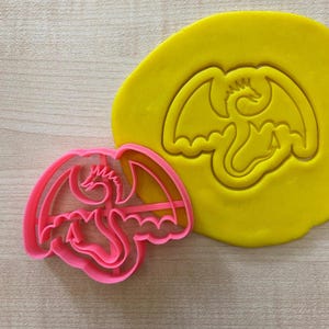 Op de afbeelding: Roze plastic koekjesvormpje in de vorm van een draak met uitgespreide vleugels. De vormpje ligt op een gele stuk modelleerklei met een afdruk van de vormpje.
