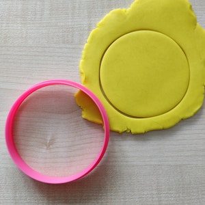 Könnte beinhalten: Ein runder Keksausstecher aus rosa Plastik neben einem gelben Teigkreis, der mit dem Ausstecher ausgestochen wurde.