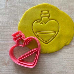 Könnte beinhalten: Rosa herzförmiger Ausstecher mit einem detaillierten Design einer herzförmigen Flasche mit einem Stopfen. Der Ausstecher wird auf einer gelben Oberfläche gezeigt.
