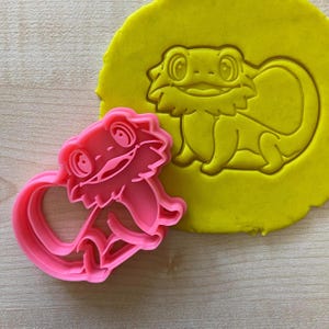 May include: Pink plastic cookie cutter shaped like a bearded dragon. The cutter is shown on top of a yellow piece of modeling clay, which has been pressed with the cutter to show the shape.