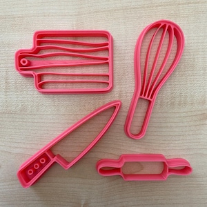 May include: Four pink plastic cookie cutters shaped like kitchen tools: a rolling pin, a knife, a chopping board, and a whisk.