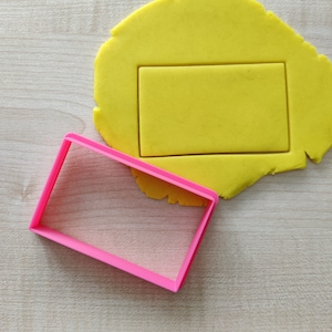 Può includere: Un tagliapasta rettangolare in plastica rosa. Il tagliapasta è sopra un pezzo di pasta frolla gialla.