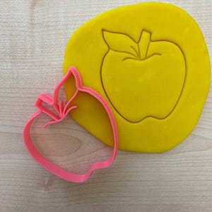 Può includere: Un tagliabiscotti rosa a forma di mela accanto a un pezzo di pasta gialla con un'impronta di mela. Il tagliabiscotti ha un design a foglia dettagliato ed è posizionato su una superficie a venatura di legno chiaro. L'impasto mostra la forma della mela.