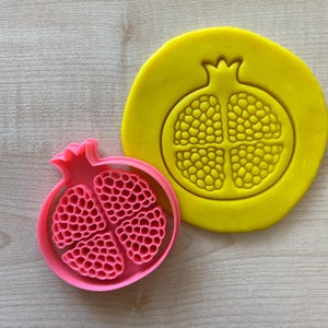 Può includere: Un tagliapasta in plastica rosa a forma di melograno, con un tappetino di impressione in plastica gialla che mostra lo stesso design.