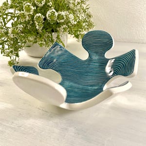 Könnte beinhalten: Eine weiße Keramikschale in Form eines Puzzlesteins mit einem blaugrünen Innenraum. Die Schale hat einen weißen Rand und ist mit dünnen weißen Linien auf dem blaugrünen Innenraum verziert.