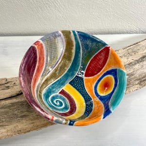 Può includere: Una piccola ciotola in ceramica colorata con un design astratto a spirale in tonalità di rosso, arancio, giallo, blu, verde e marrone.