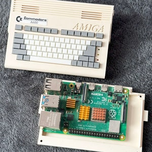 Puede incluir: Un ordenador Commodore A600 beige con un teclado y la palabra "AMIGA" impresa en la parte superior. Una placa de ordenador Raspberry Pi 4 Model B está sentada dentro de una caja blanca.