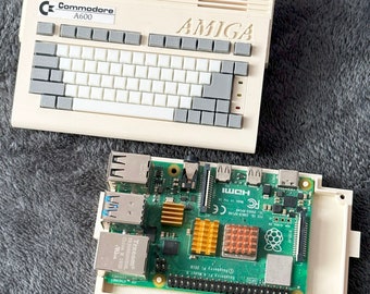 Peut inclure: Un ordinateur Commodore A600 beige avec un clavier et le mot "AMIGA" imprimé sur le dessus. Une carte d'ordinateur Raspberry Pi 4 Model B est placée dans un boîtier blanc.