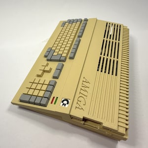 以下が含まれることがあります： ベージュのAmigaコンピュータで、キーボードと換気口があります。コンピュータには黒と緑のロゴがあり、側面に「AMIGA」と印刷されています。