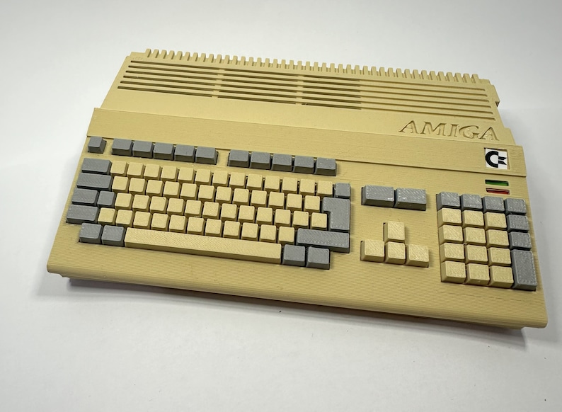 以下が含まれることがあります： ベージュの Amiga コンピューター。グレーのキーボードと、上部に「AMIGA」という文字が印刷されています。コンピューターにはコモドールロゴとイタリア国旗があります。