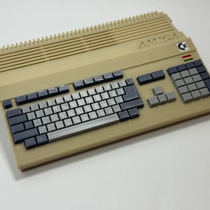 Op de afbeelding: Een beige Amiga-computer met een grijs toetsenbord en een grijs numeriek toetsenblok. De computer heeft een zwart-witte sticker met het woord "AMIGA" en een kleine, kleurrijke sticker met een "C" erop.