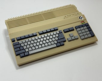 Peut inclure: Un ordinateur Amiga beige avec un clavier gris et un pavé numérique gris. L'ordinateur a un autocollant noir et blanc avec le mot "AMIGA" et un petit autocollant coloré avec un "C" dessus.