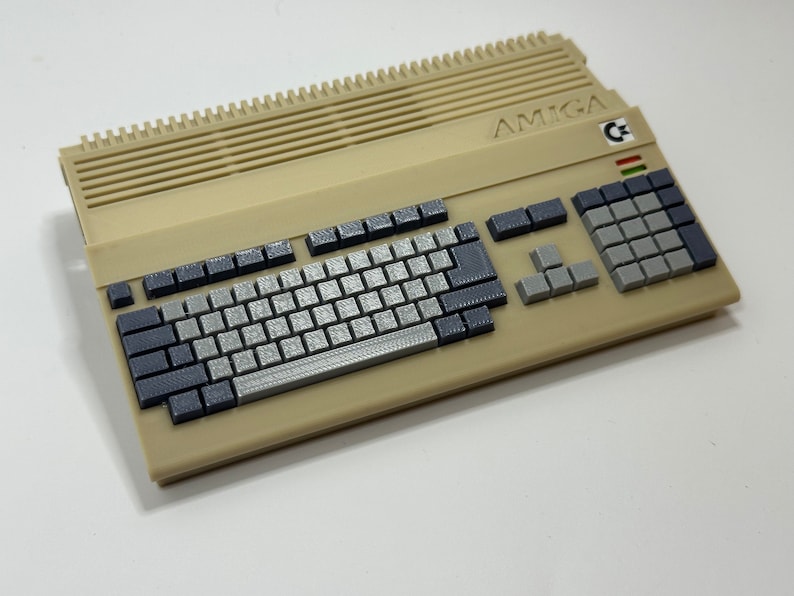 以下が含まれることがあります： グレーのキーボードとグレーのテンキーが付いたベージュのAmigaコンピューター。コンピューターには、「AMIGA」という文字の白黒のステッカーと、「C」のカラフルな小さなステッカーが付いています。