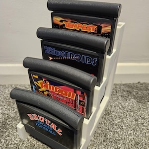Könnte beinhalten: Ein weißer Videospiel-Kassettenhalter aus Kunststoff mit vier schwarzen Kassetten. Die Kassetten sind mit "TEMPEST", "REBOOTEROIDS", "PINBALL" und "BRUTAL SPORTS FOOTBALL" beschriftet. Der Halter steht auf einem grauen Teppich.