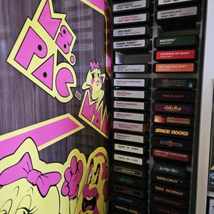 以下が含まれることがあります： Atari 7800 および 2600 のビンテージビデオゲームカートリッジのディスプレイ。 ピンクと黄色のパックマンのキャラクターとロゴが特徴のパックマンをテーマにしたデザイン。 黒いカートリッジにはゲームタイトルが印刷されています。