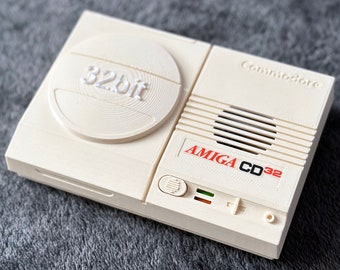 Peut inclure: Une console Commodore Amiga CD32 blanche avec un logo 32 bits sur le devant. La console possède un lecteur de CD, un haut-parleur et un bouton d'alimentation.