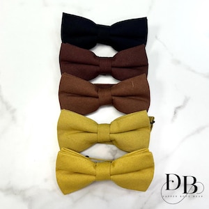 Può includere: Quattro papillon in fila, ognuno di un colore diverso. Il papillon in alto è nero, seguito da due tonalità di marrone, e i due inferiori sono gialli. I papillon sono realizzati in tessuto strutturato.