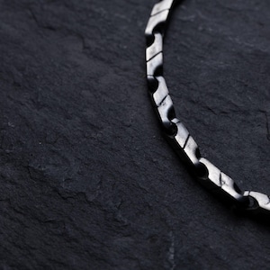 May include: Close-up of a silver-colored chain necklace with a geometric design. The necklace is made of interlocking, angular links. The background is a dark, textured surface, providing contrast to the metallic chain.