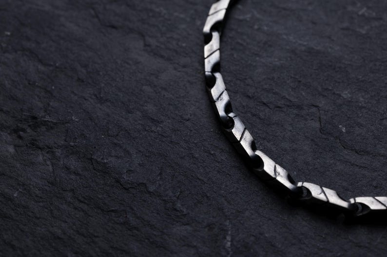 May include: Close-up of a silver-colored chain necklace with a geometric design. The necklace is made of interlocking, angular links. The background is a dark, textured surface, providing contrast to the metallic chain.