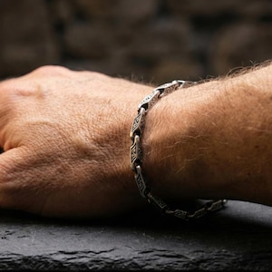 May include: Silver-toned bracelet with an intricate, repeating geometric design. The bracelet is worn on a wrist, with the hand resting on a dark surface. The bracelet's links are detailed with patterns.