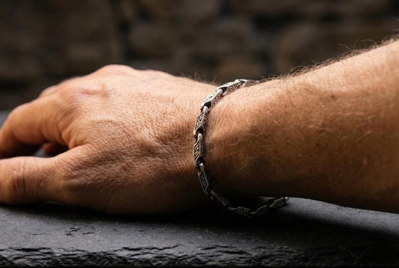 May include: Silver-toned bracelet with an intricate, repeating geometric design. The bracelet is worn on a wrist, with the hand resting on a dark surface. The bracelet's links are detailed with patterns.