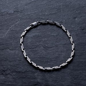 May include: A silver bracelet with a repeating geometric design. The bracelet is made of linked segments, each with a diamond-shaped pattern. The clasp is visible. The bracelet is displayed on a dark, textured surface.