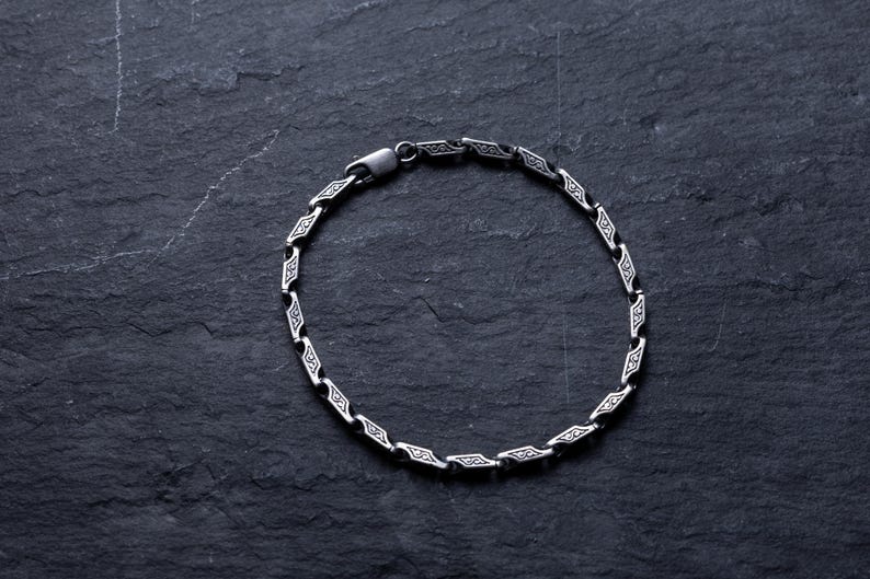 May include: A silver bracelet with a repeating geometric design. The bracelet is made of linked segments, each with a diamond-shaped pattern. The clasp is visible. The bracelet is displayed on a dark, textured surface.