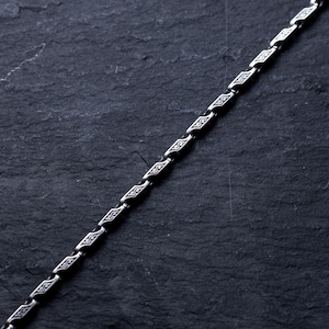 May include: A silver chain with a repeating geometric pattern. The chain is angled across a dark, textured surface. The links have a diamond-like shape with etched details, creating a decorative and elegant appearance. The chain is likely for a necklace or bracelet.