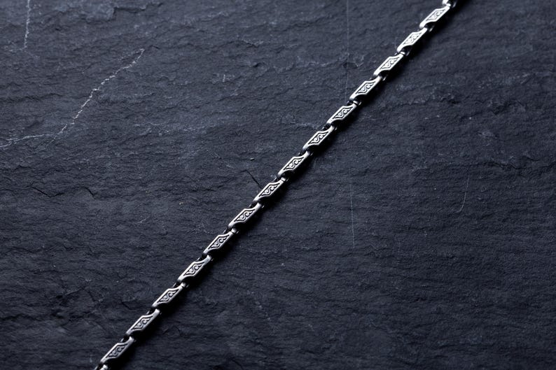 May include: A silver chain with a repeating geometric pattern. The chain is angled across a dark, textured surface. The links have a diamond-like shape with etched details, creating a decorative and elegant appearance. The chain is likely for a necklace or bracelet.