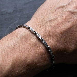 May include: Silver bracelet featuring a detailed, repeating pattern. The bracelet is composed of linked segments, each with an intricate design. The bracelet is worn on a wrist, highlighting the detailed metalwork. The bracelet is a piece of jewellery.