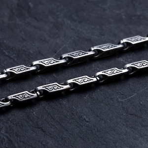 May include: Close-up of two silver-colored chains with a repeating geometric design. Each link features an engraved pattern. The chains are displayed on a dark, textured surface, creating a contrast that highlights the intricate details of the jewelry.