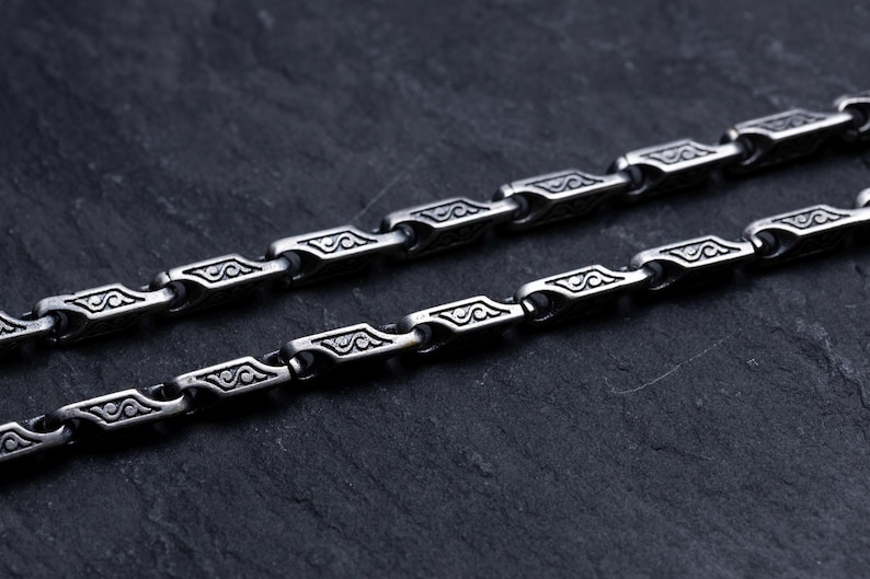 May include: Close-up of two silver-colored chains with a repeating geometric design. Each link features an engraved pattern. The chains are displayed on a dark, textured surface, creating a contrast that highlights the intricate details of the jewelry.