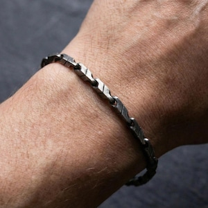 May include: A silver-toned bracelet with a geometric design. The bracelet is composed of rectangular links with etched details, connected by small, round links. The bracelet is worn on a person's wrist.