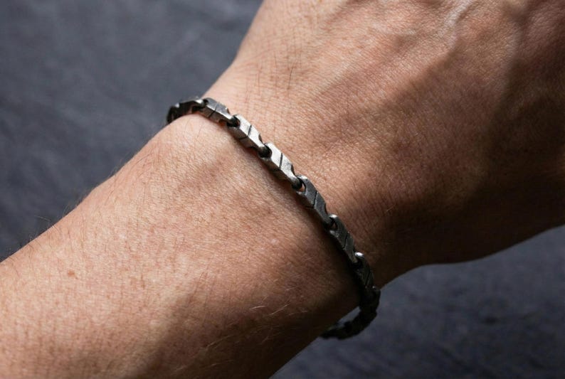 May include: A silver-toned bracelet with a geometric design. The bracelet is composed of rectangular links with etched details, connected by small, round links. The bracelet is worn on a person's wrist.