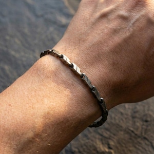 May include: A silver-toned bracelet with a geometric design is worn on a person's wrist. The bracelet is composed of interconnected, arrow-shaped links. The image is taken in natural light, highlighting the bracelet's texture and details.