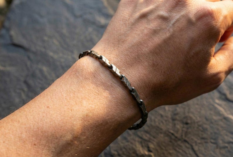 May include: A silver-toned bracelet with a geometric design is worn on a person's wrist. The bracelet is composed of interconnected, arrow-shaped links. The image is taken in natural light, highlighting the bracelet's texture and details.