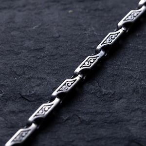 May include: Close-up of a silver chain with a repeating geometric pattern. The chain links are diamond-shaped with ornate designs. The chain is displayed against a dark, textured background, highlighting the intricate details of the jewelry.