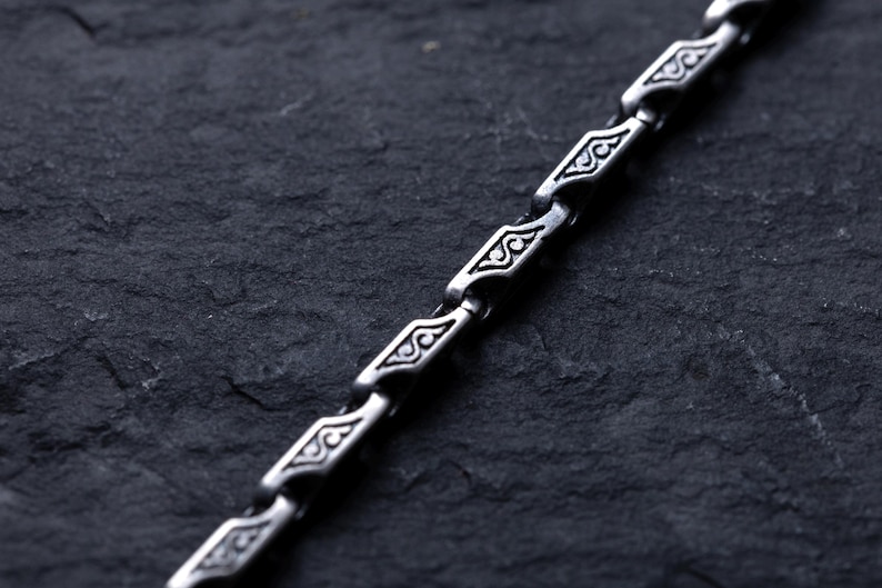 May include: Close-up of a silver chain with a repeating geometric pattern. The chain links are diamond-shaped with ornate designs. The chain is displayed against a dark, textured background, highlighting the intricate details of the jewelry.