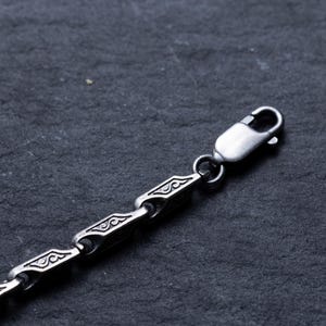 May include: Close-up of a silver-colored chain necklace with a decorative pattern. The chain is composed of rectangular links with an engraved design, connected by smaller, rounded links. A lobster clasp secures the necklace.