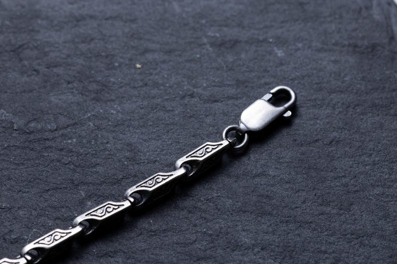 May include: Close-up of a silver-colored chain necklace with a decorative pattern. The chain is composed of rectangular links with an engraved design, connected by smaller, rounded links. A lobster clasp secures the necklace.