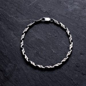May include: A silver bracelet with a unique, textured link design. The bracelet is made of a shiny, metallic material and features a secure clasp. The jewelry is displayed on a dark, textured surface.