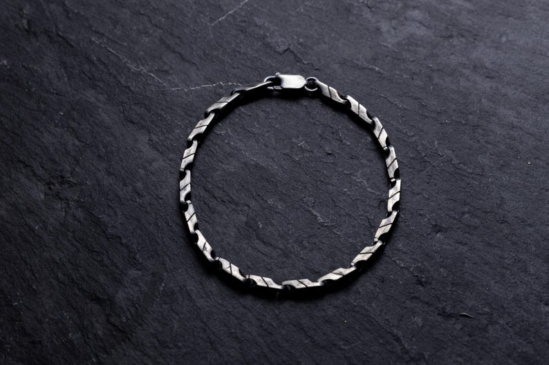 May include: A silver bracelet with a unique, textured link design. The bracelet is made of a shiny, metallic material and features a secure clasp. The jewelry is displayed on a dark, textured surface.