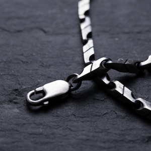 May include: Close-up of a silver-toned necklace with a unique, geometric link design. The necklace features a lobster clasp closure. The jewelry is displayed against a dark, textured background, highlighting the metallic sheen of the chain.