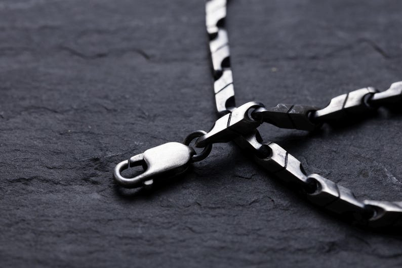 May include: Close-up of a silver-toned necklace with a unique, geometric link design. The necklace features a lobster clasp closure. The jewelry is displayed against a dark, textured background, highlighting the metallic sheen of the chain.