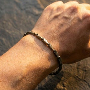 May include: A silver-toned bracelet with a geometric design is worn on a wrist. The bracelet is composed of interconnected, arrow-shaped links. The skin tone is light, and the background is a blurred, natural setting.